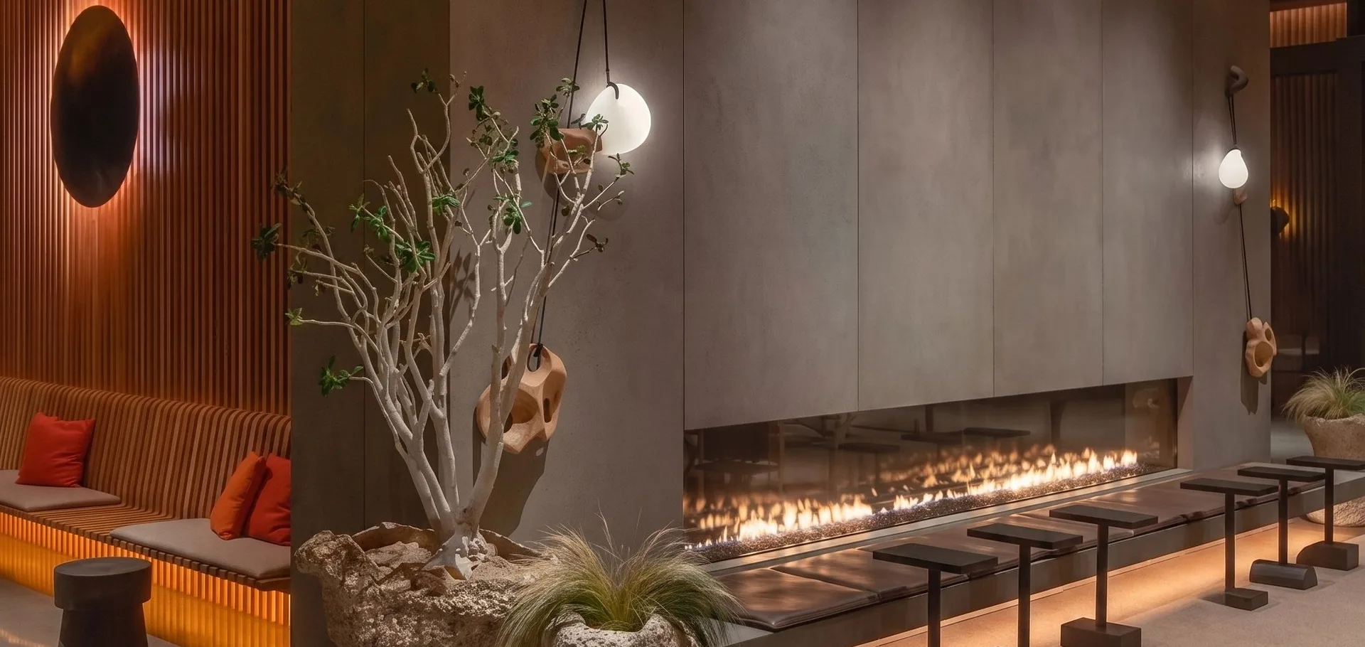A long linear gas fireplace running the length of a concrete feature wall in a hotel lobby, with banquette seating and bar stools along it