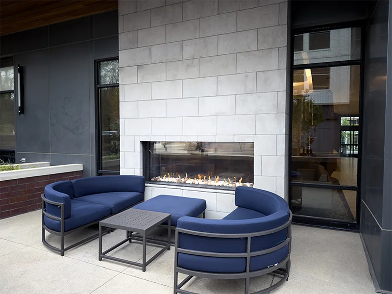 An outdoor see-through gas fireplace in a light block surround on a commercial patio, with curved blue lounge seating around it