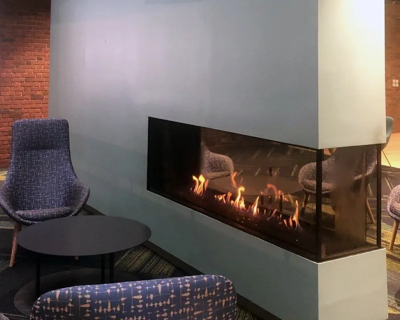 A large see-through gas fireplace set into a freestanding feature wall in a commercial lounge, with lounge chairs visible through the fire from the other side