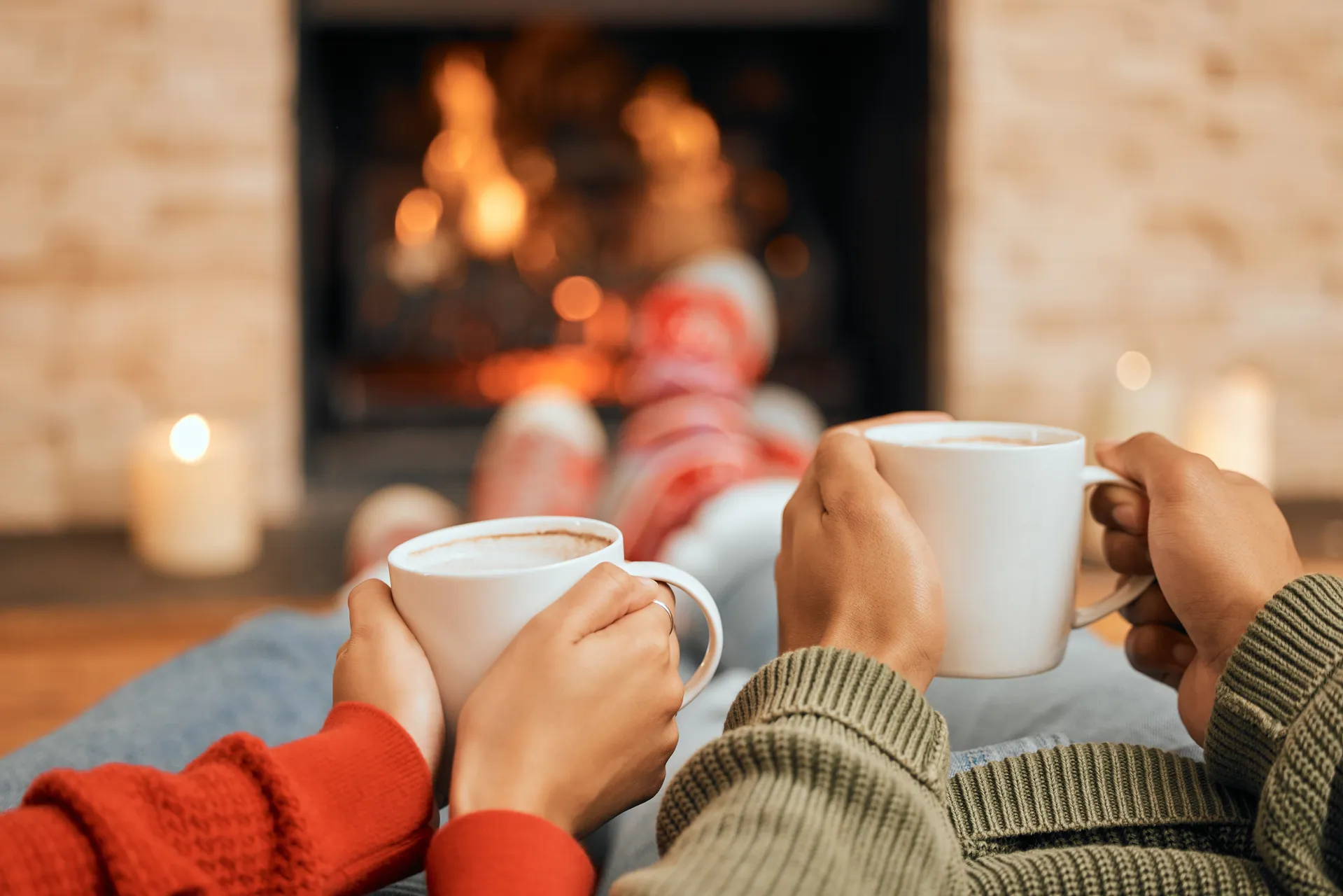 Two people with feet up by a lit fireplace, holding mugs
