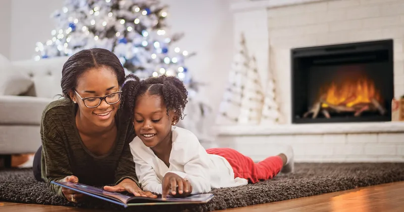 A woman and a girl reading on the rug in front of a lit electric fireplace