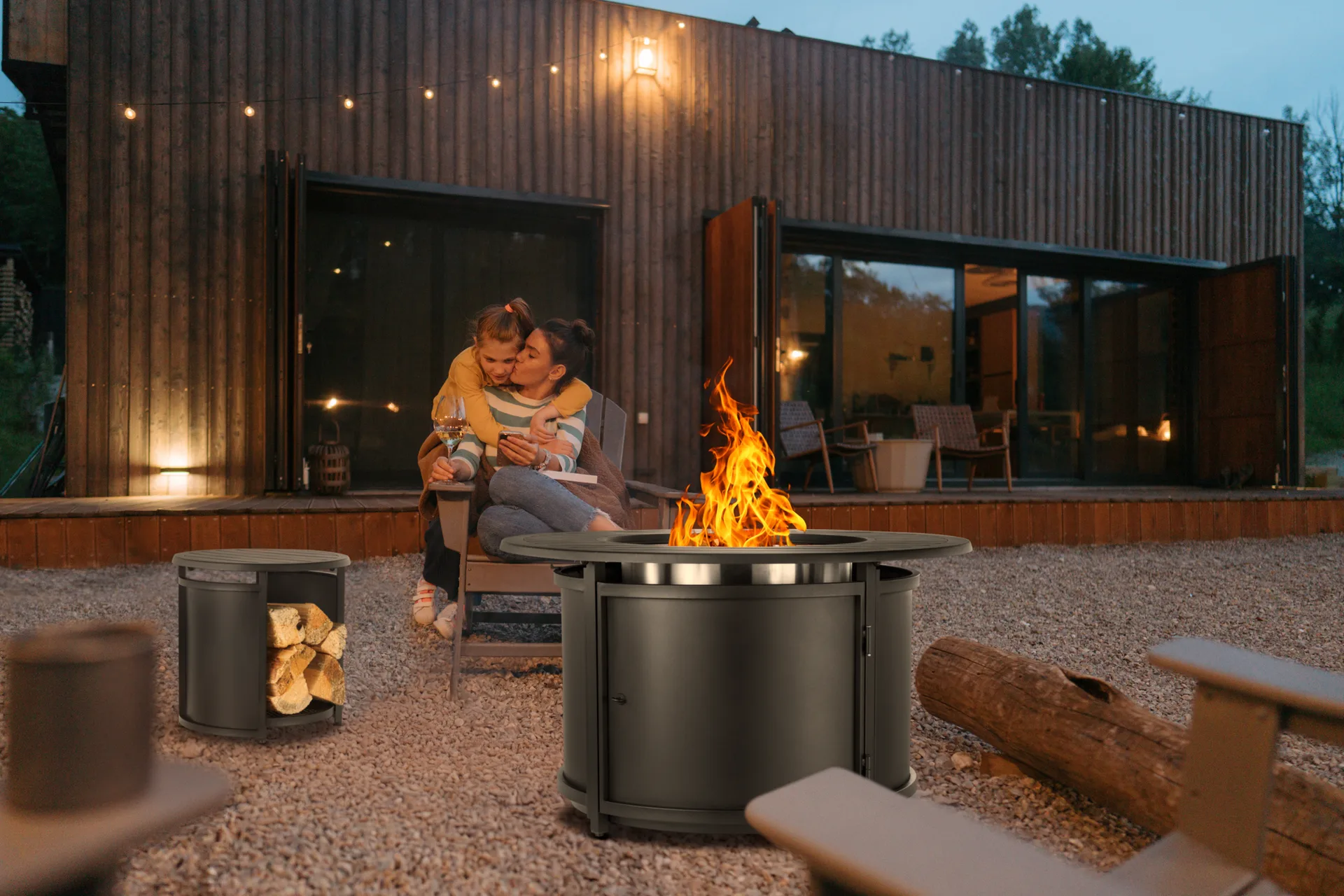 Mother and child by a lit fire pit at dusk outside a cedar-clad home