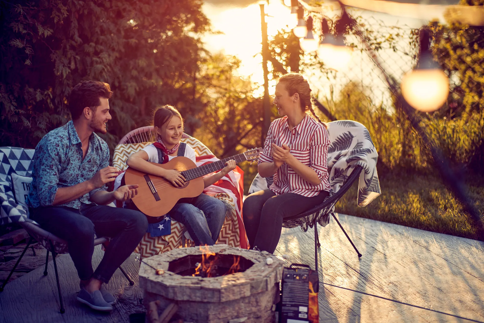 Family around a backyard fire pit at sunset, one playing guitar