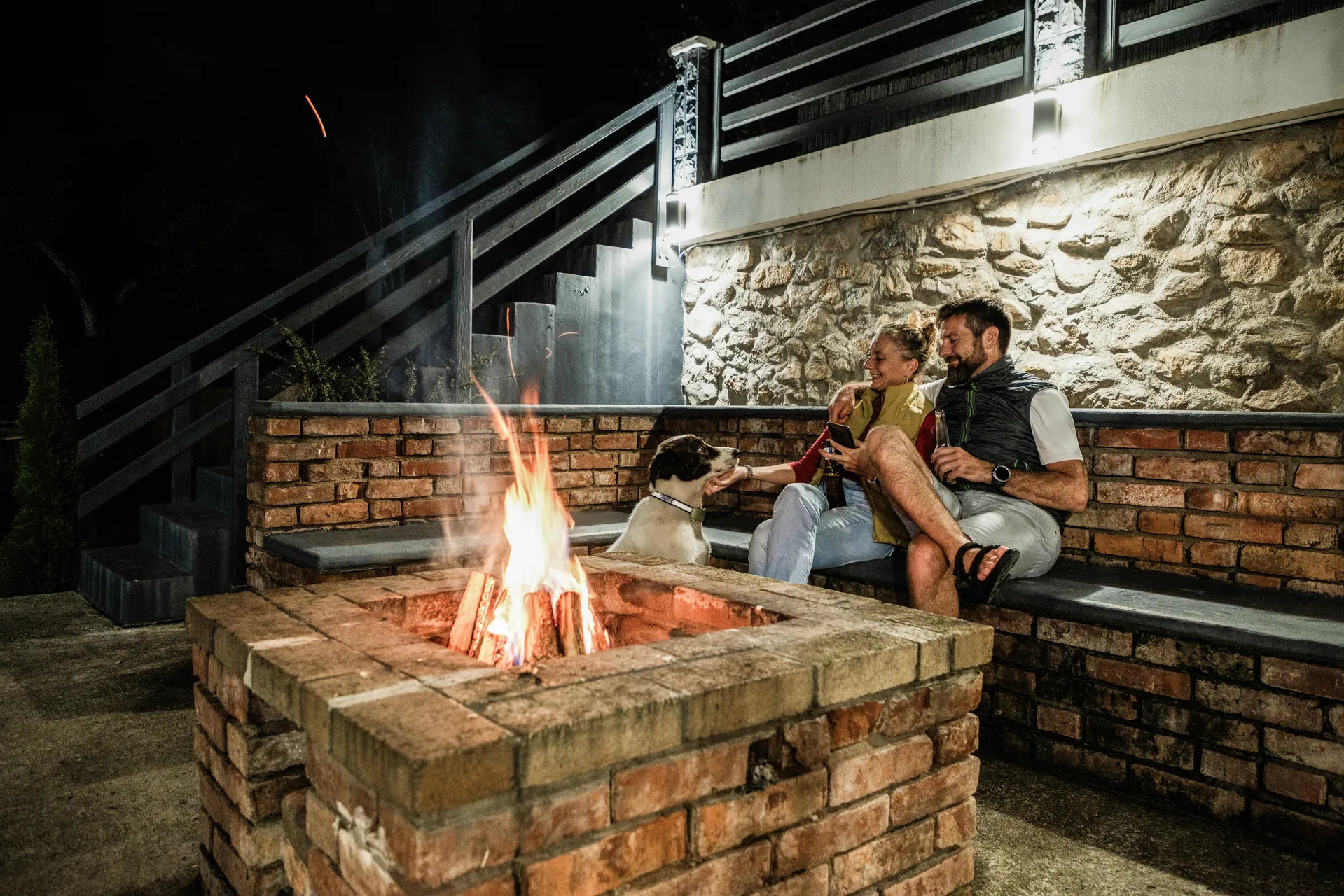 Family at a brick fire pit at night beside a modern home