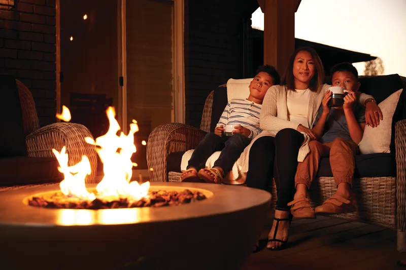 A family of four on a covered porch behind a lit round fire table at night