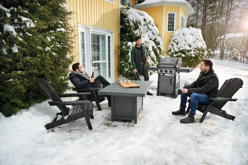 Three men in Adirondack chairs around a lit fire table in a snowy yard