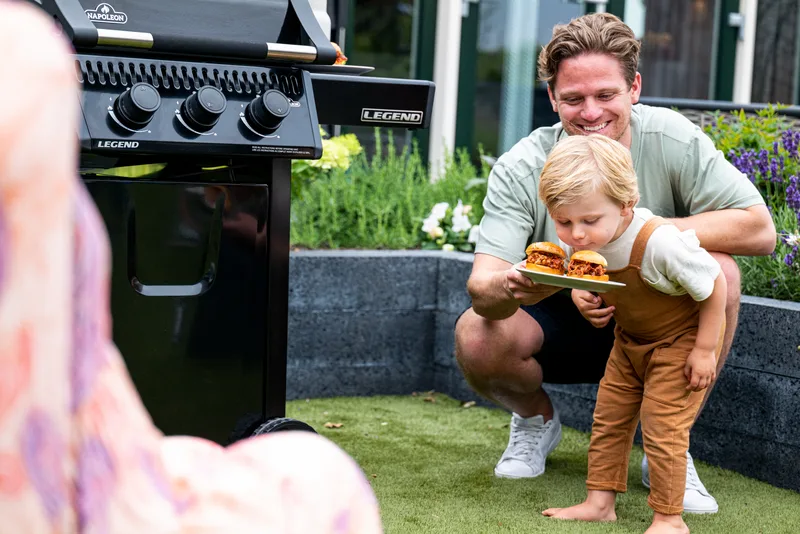 A father crouching with a toddler holding a plate at a gas grill